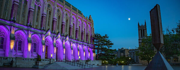 Suzzallo Library at the University of Washington Seattle campus at night is lit by purple floodlights, with a sculpture in the foreground and the moon beyond.